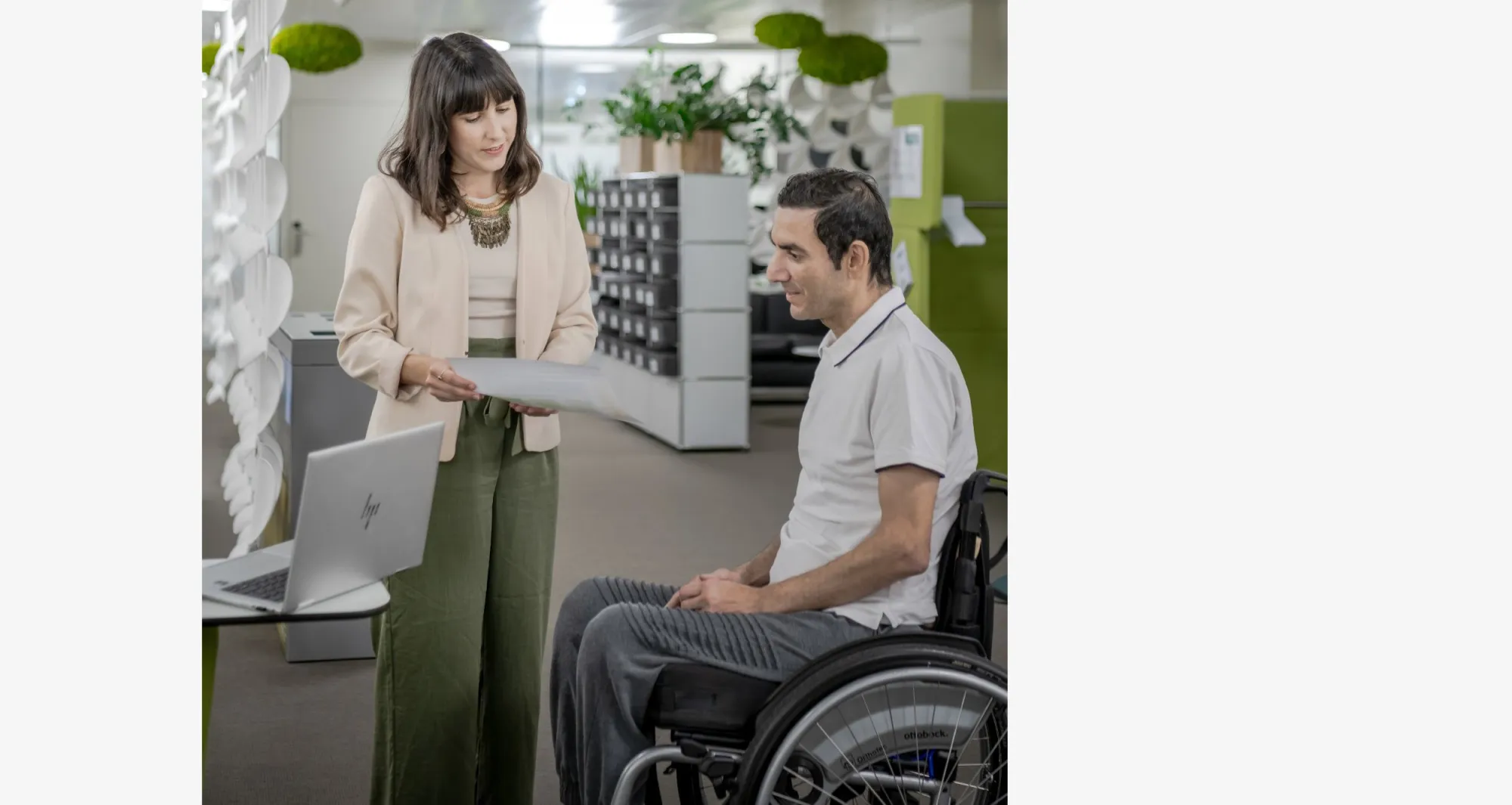 In einem modernen Büro sind zwei Personen in einem Gespräch. Die Frau links, Selina Infanger trägt eine helle Jacke, ein helles Oberteil und grüne Hosen. Sie steht, hält ein Dokument in der Hand und liest etwas darauf vor. Der Mann rechts sitzt in einem Rollstuhl und trägt ein weißes T-Shirt und graue Hosen. Vor ihnen auf einem kleinen Tisch steht ein aufgeklappter Laptop. Der Hintergrund zeigt eine offene Büroeinrichtung mit Pflanzen und modernen Dekorationselementen wie hängenden grünen Moosobjekten.
