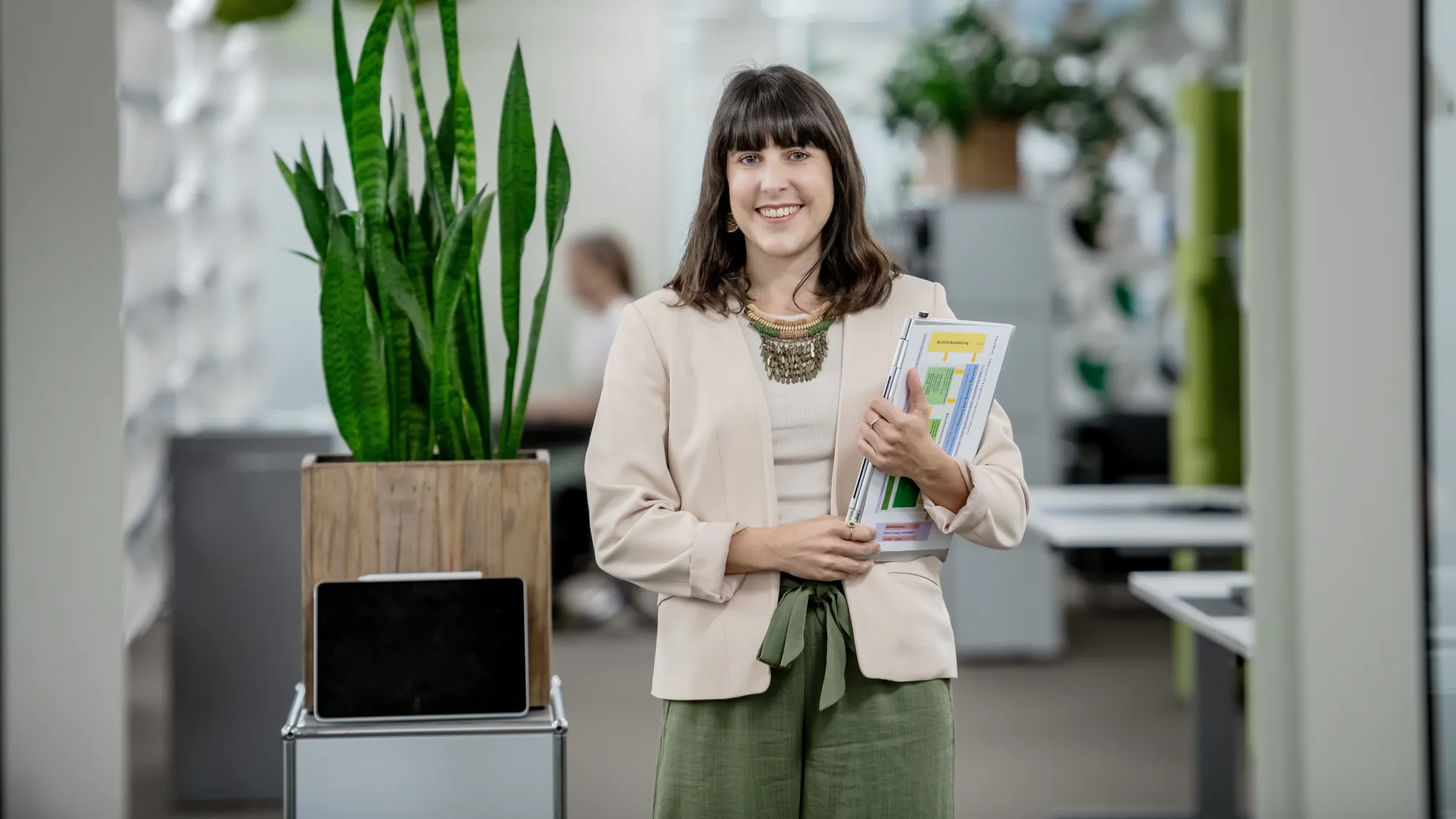 Selina Infanger steht lächelnd in einem modernen Büro. Sie trägt eine helle Jacke, ein beigefarbenes Oberteil und weite grüne Hosen. In ihren Armen hält sie mehrere Dokumente und Unterlagen. Im Hintergrund befinden sich Büropflanzen, darunter eine hohe Grünpflanze in einem Holztopf, der auf einem kleinen Regal steht, sowie weitere Dekorationselemente und Möbel. Die Büroeinrichtung wirkt hell und offen, mit modernen Gestaltungselementen und einer klaren, strukturierten Umgebung. 