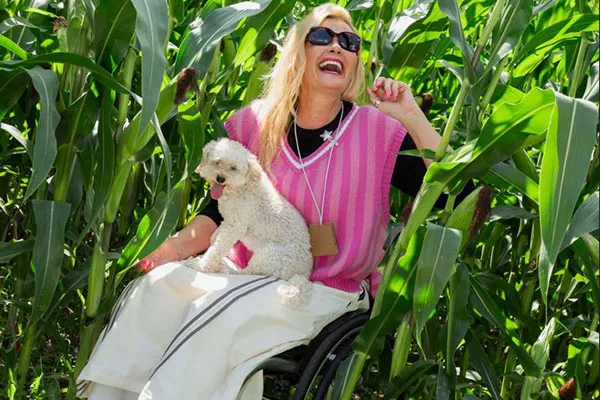 Rollstuhlfahrerin sitzt lachend in einem Maisfeld mit einem kleinen Hund auf dem Schoss.
